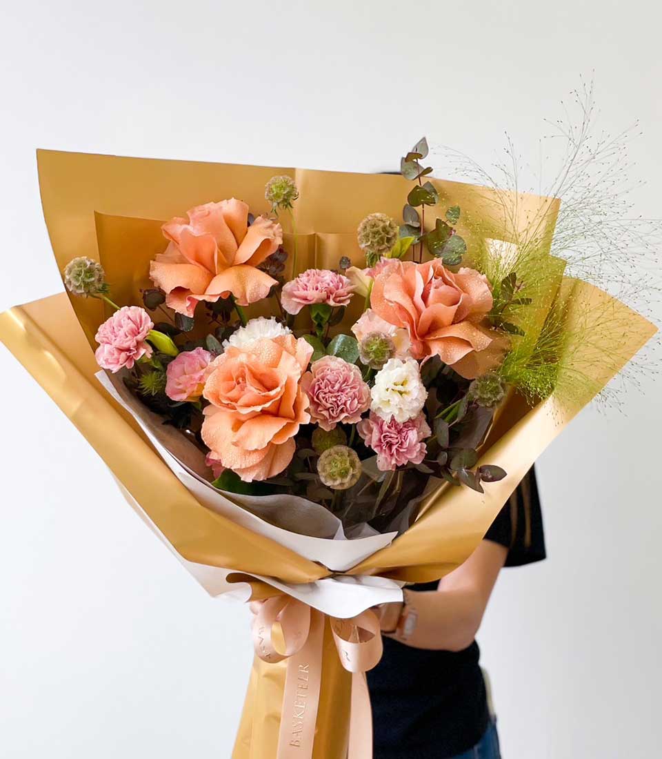 Shimmering peach rose bouquet with pink and white carnations, wrapped in gold paper with pink ribbon, perfect for graduation