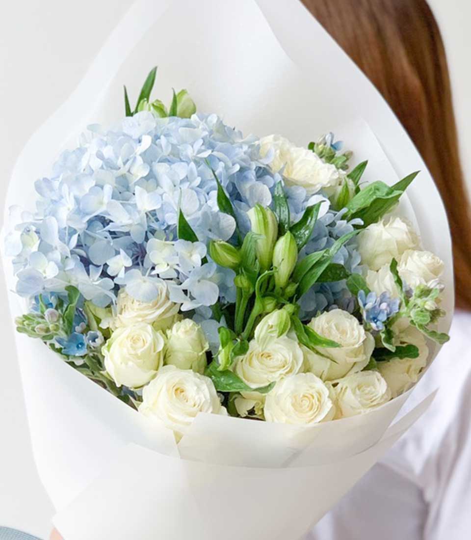 Fresh blue hydrangea and white rose bouquet with green foliage, wrapped in white paper for birthdays and celebrations