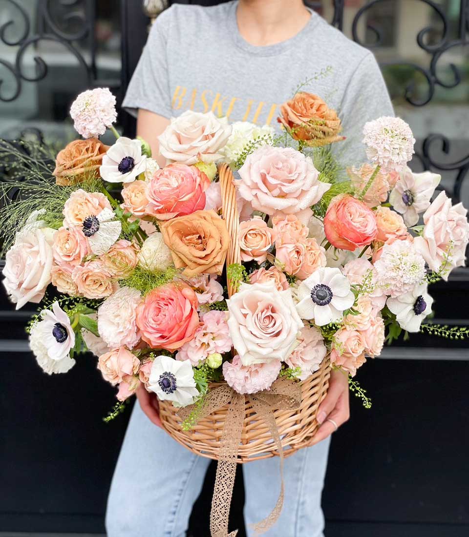 Overflowing wicker basket of fresh pink, peach, and coral roses with white anemones and lush green foliage