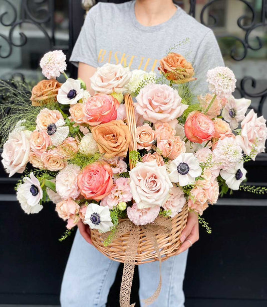 Overflowing wicker basket of fresh pink, peach, and coral roses with white anemones and lush green foliage