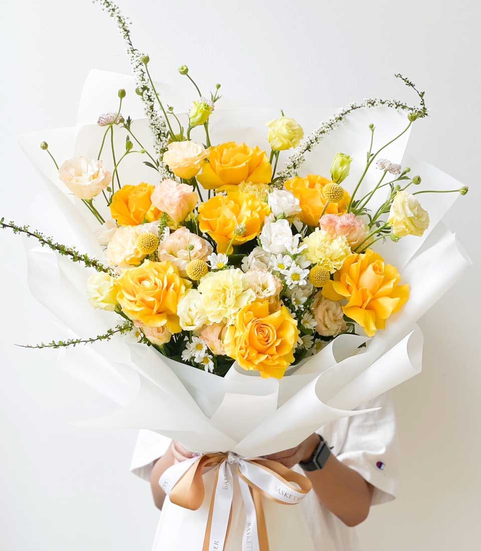 Bright yellow and peach fresh flower bouquet with roses, carnations, and daisies, wrapped in white paper with gold ribbon