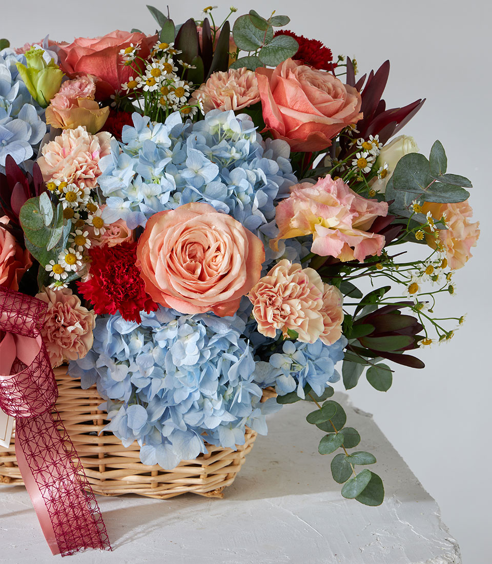 Vibrant get well flowers basket with blue hydrangeas, peach roses, and pink carnations in woven wicker