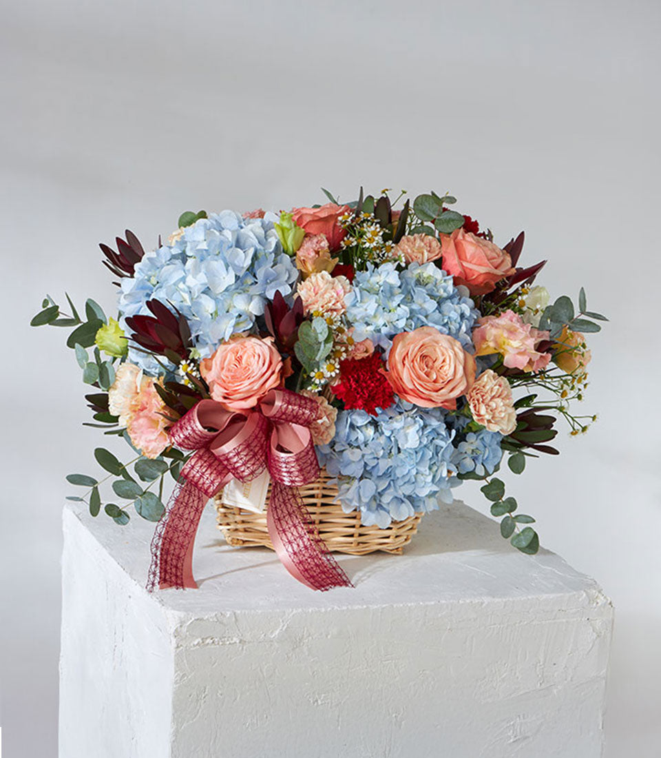 Bright multicoloured fresh flower basket with hydrangeas, roses, and carnations in woven basket with burgundy ribbon