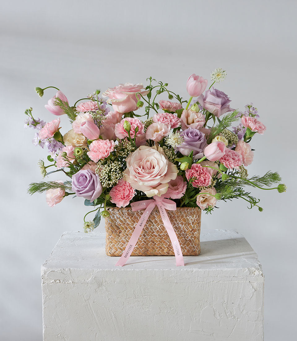 Lush lavender and pink rose arrangement in woven basket with cream filler flowers and green foliage
