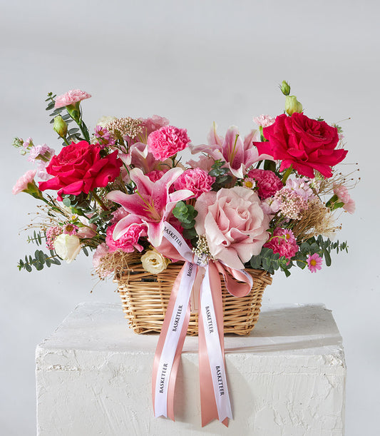Pink and fuchsia roses with lilies in woven basket, birthday flowers with white ribbon accent