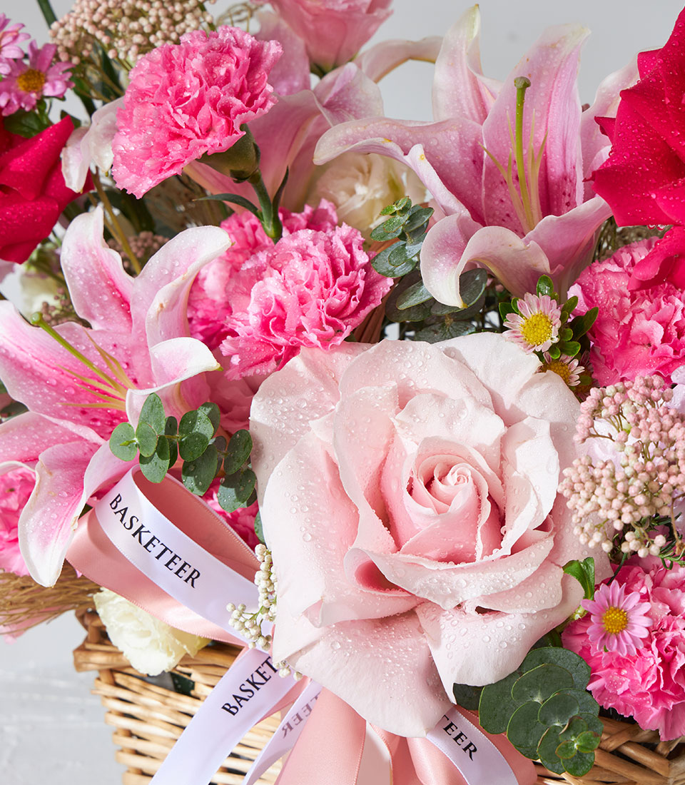 Pink roses, lilies, and carnations in woven basket with eucalyptus and ribbon, birthday flower gift