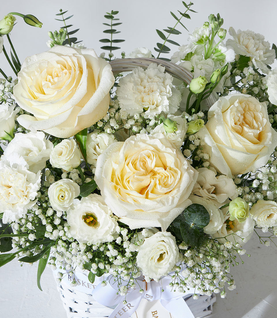 White roses, eustoma, and baby's breath arrangement in woven basket with eucalyptus foliage