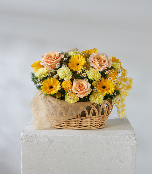 Blissful Harmony bouquet with yellow gerberas, peach roses, and carnations in woven wicker basket with gingham ribbon