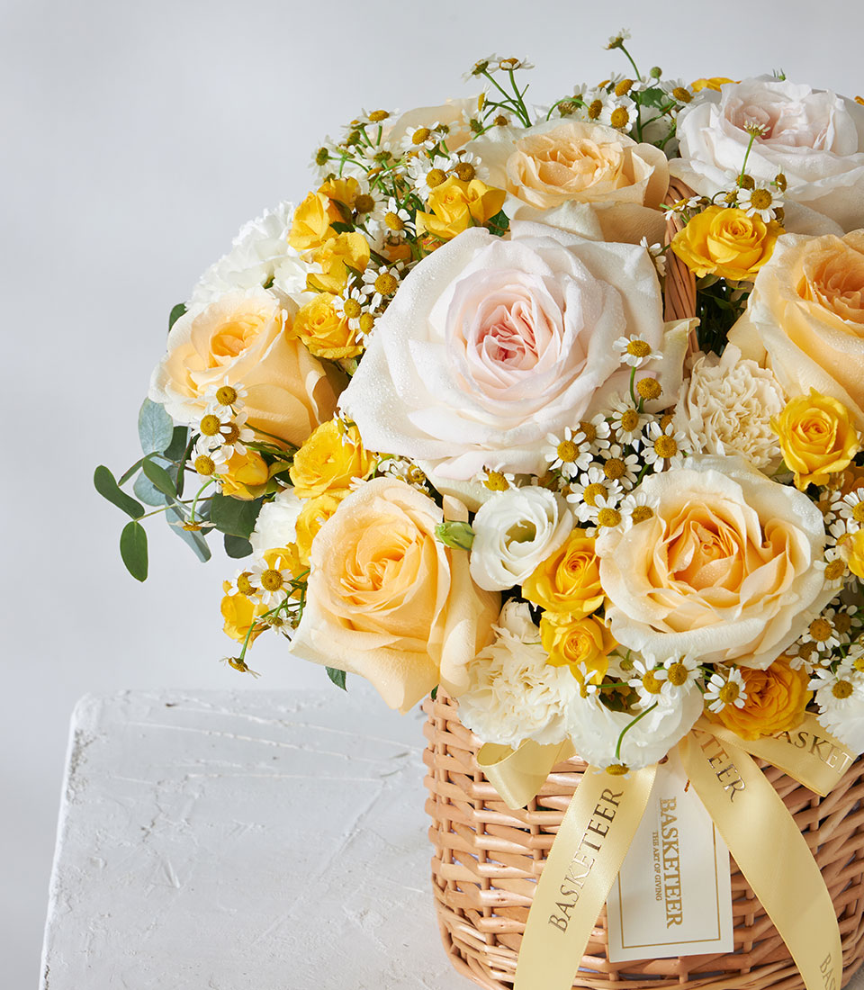 Pale peach, cream, and yellow roses with white daisies in a woven wicker basket, tied with golden ribbon