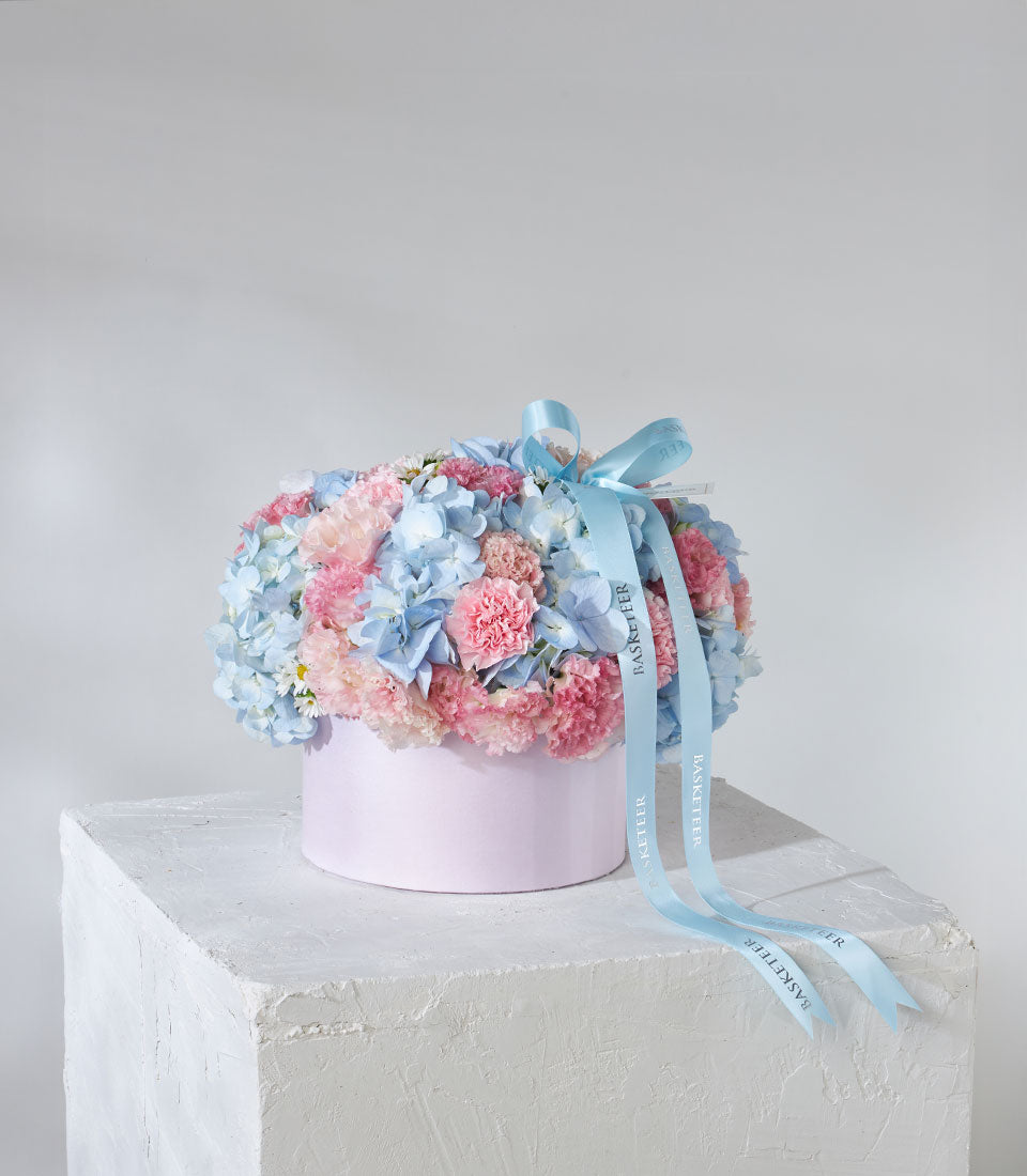 Pastel blue hydrangeas and pink carnations in light pink hatbox with ribbon