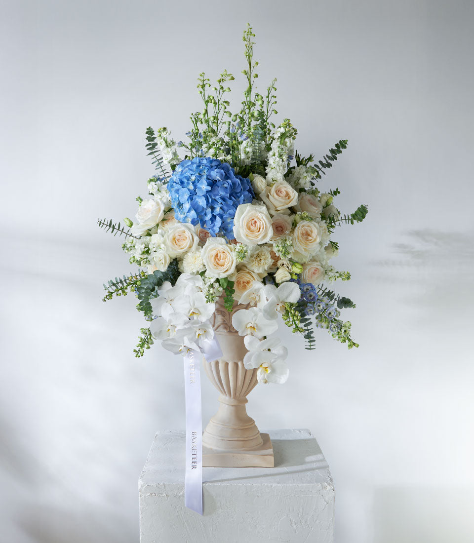 White O'Hara roses and blue hydrangeas in classical pedestal vase with eucalyptus and orchids