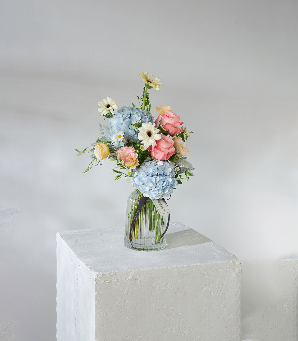 Light blue hydrangeas and pink roses in clear glass vase with seasonal blooms and green foliage