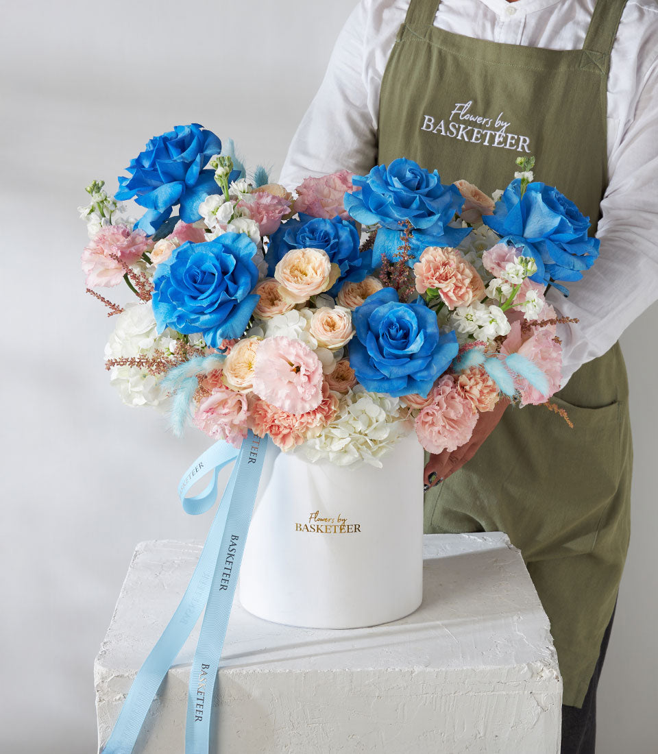 Florist holding premium blue rose and pastel garden rose arrangement in white box with blue ribbon