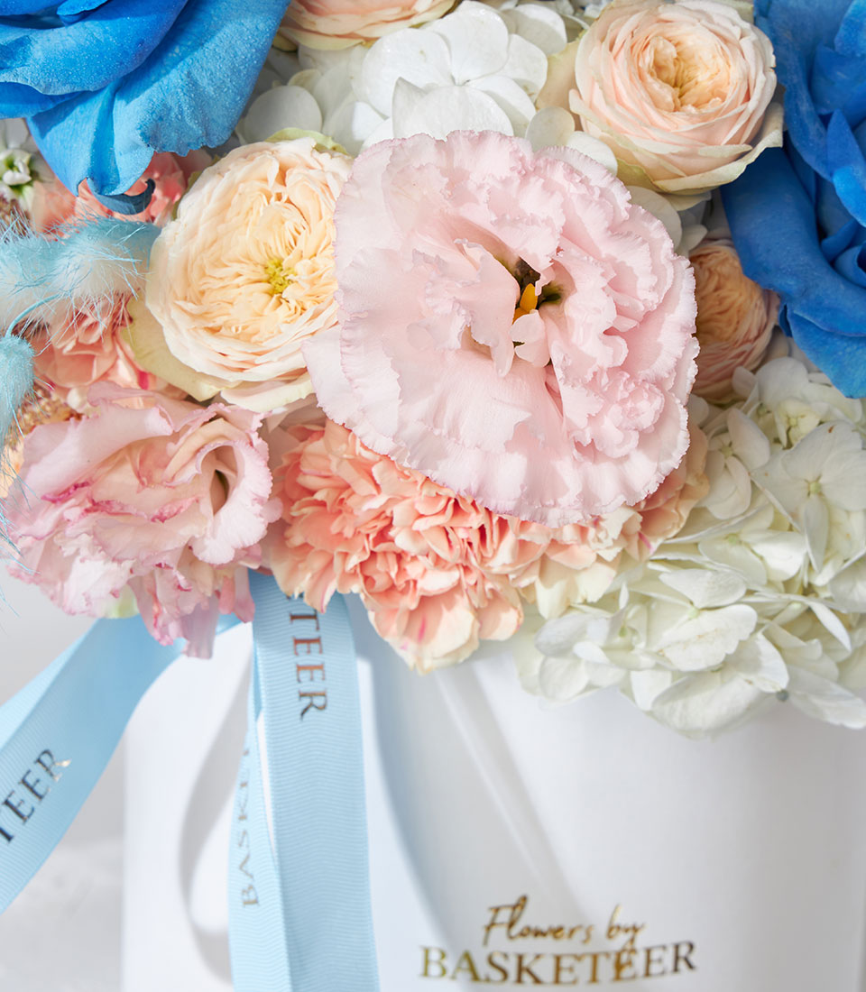 Pastel pink roses, peach garden roses, and blue-tinted rose in white cylinder box with blue ribbon
