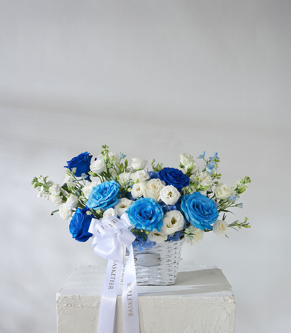 Tinted blue roses and white hydrangea arrangement in white wicker basket with satin ribbon