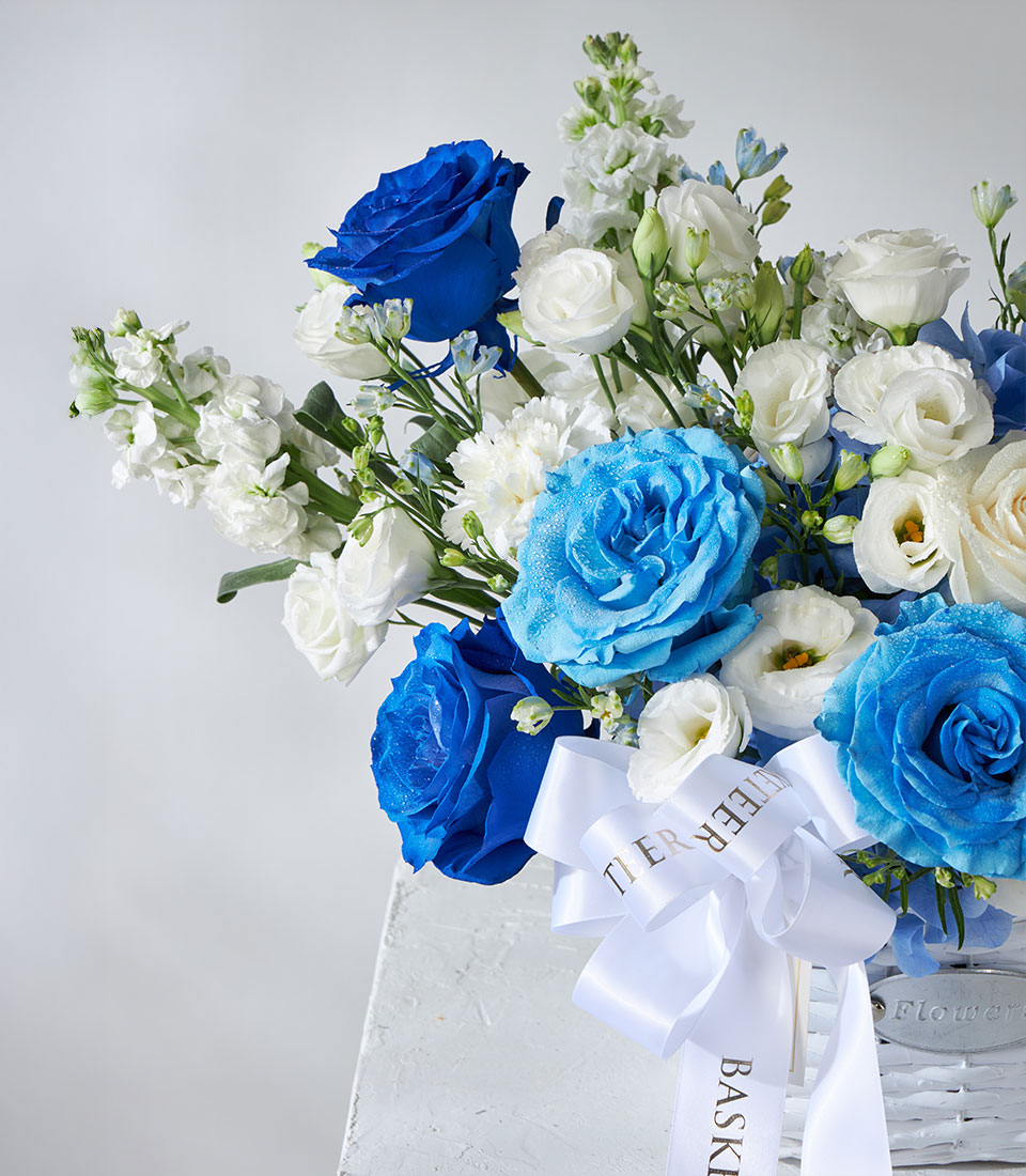Tinted blue roses and white hydrangea arrangement in white wicker basket with satin ribbon