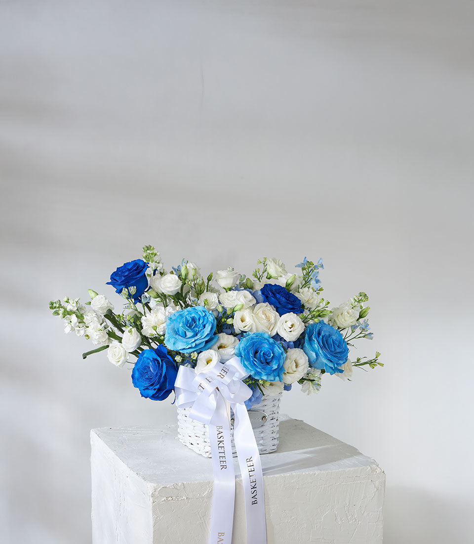 Tinted blue roses and white hydrangea in woven basket with satin ribbon