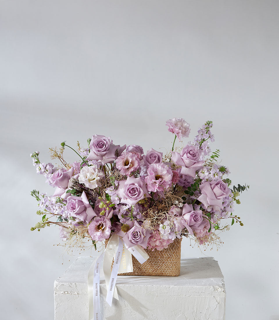 Lush purple roses and lavender hydrangea arrangement in woven basket with fresh filler flowers and greenery