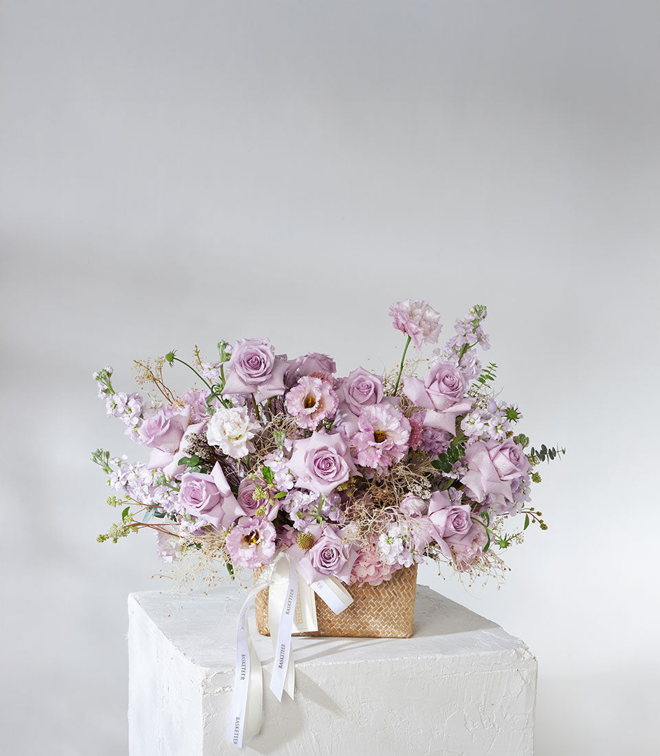 Lush purple and lavender roses with hydrangea in natural woven gift basket, tied with white ribbons