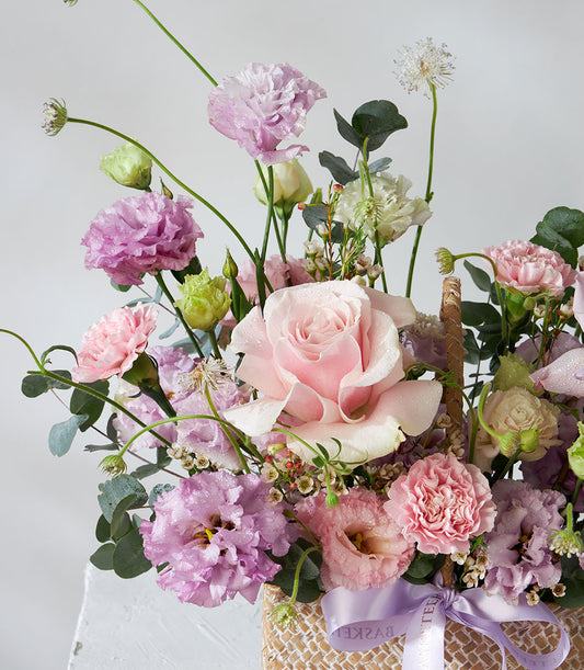 Woven basket arrangement with pink roses, purple lisianthus, and white filler flowers tied with satin ribbon