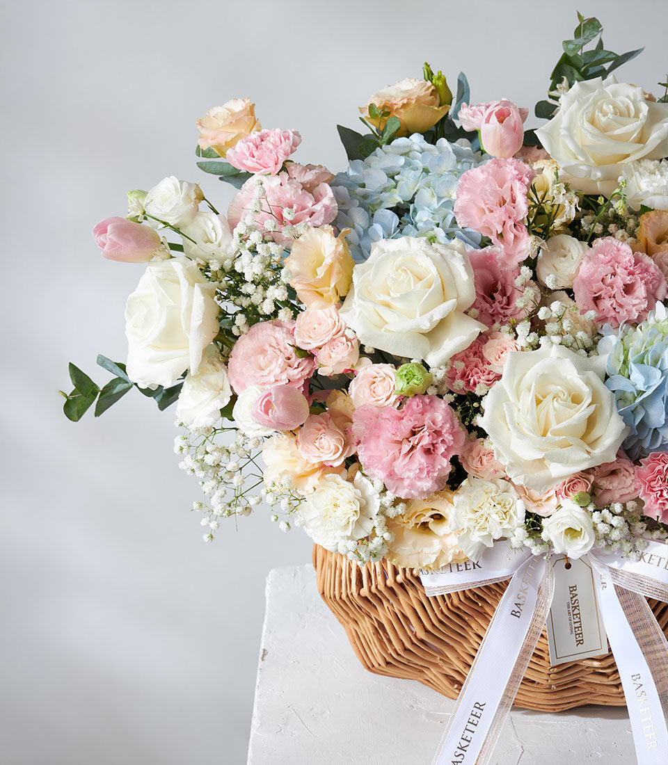 Elegant flower arrangement by Flowers by Basketeer. White roses, pink carnations, and blue hydrangeas fill a woven basket. Premium flowers for flower delivery Bangkok.