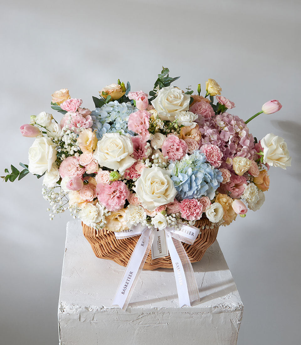 Pastel fresh flower arrangement in wicker basket with white roses, pink carnations, and blue hydrangeas for birthdays and