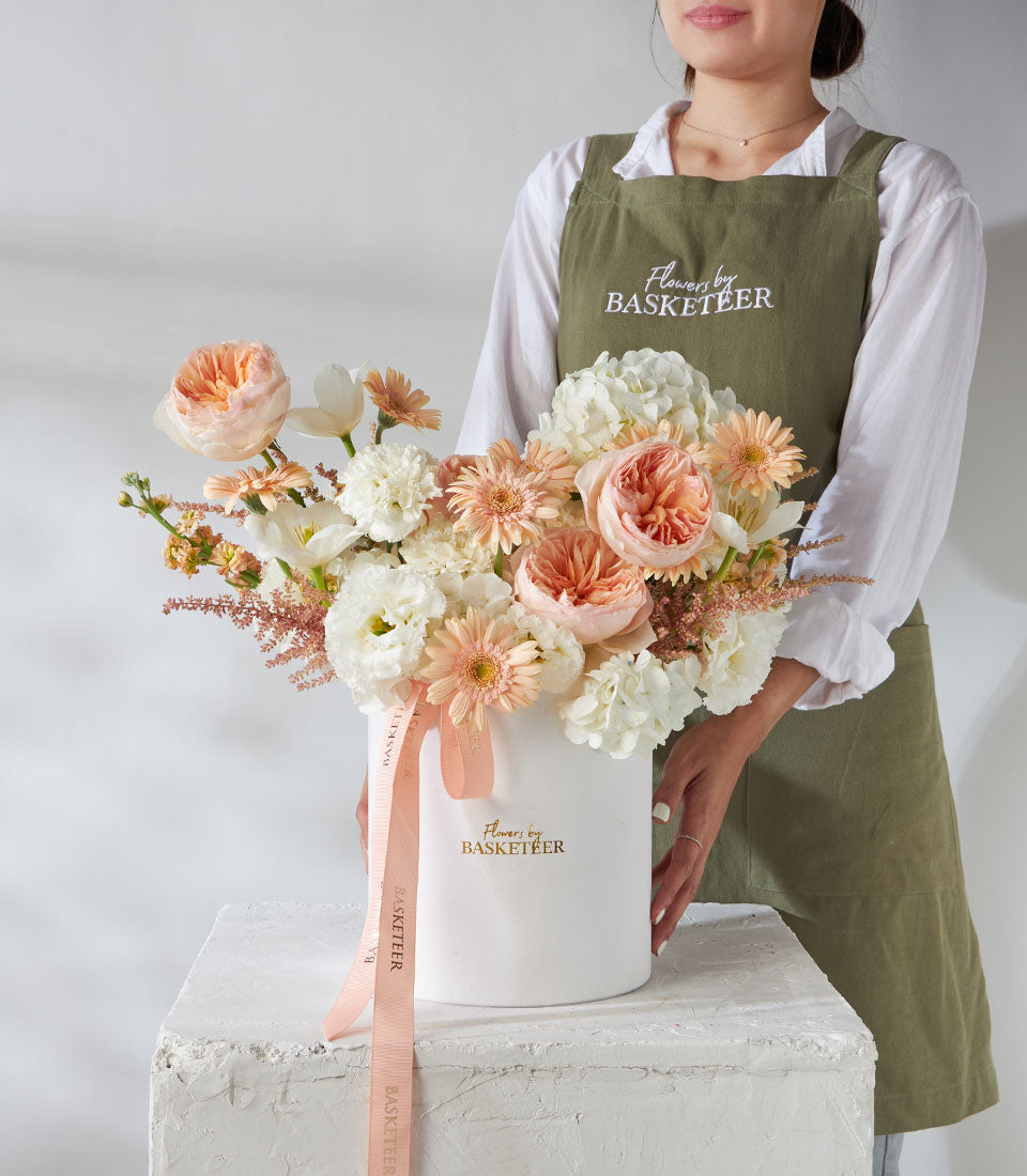 Luxury cylindrical flower box with white, peach, and coral roses, gerberas, and hydrangeas tied with peach ribbon