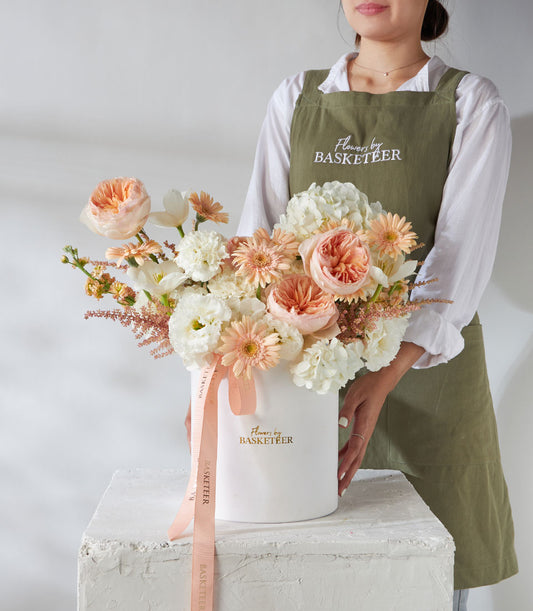 Luxury cylindrical flower box with white, peach, and coral roses, gerberas, and hydrangeas tied with peach ribbon