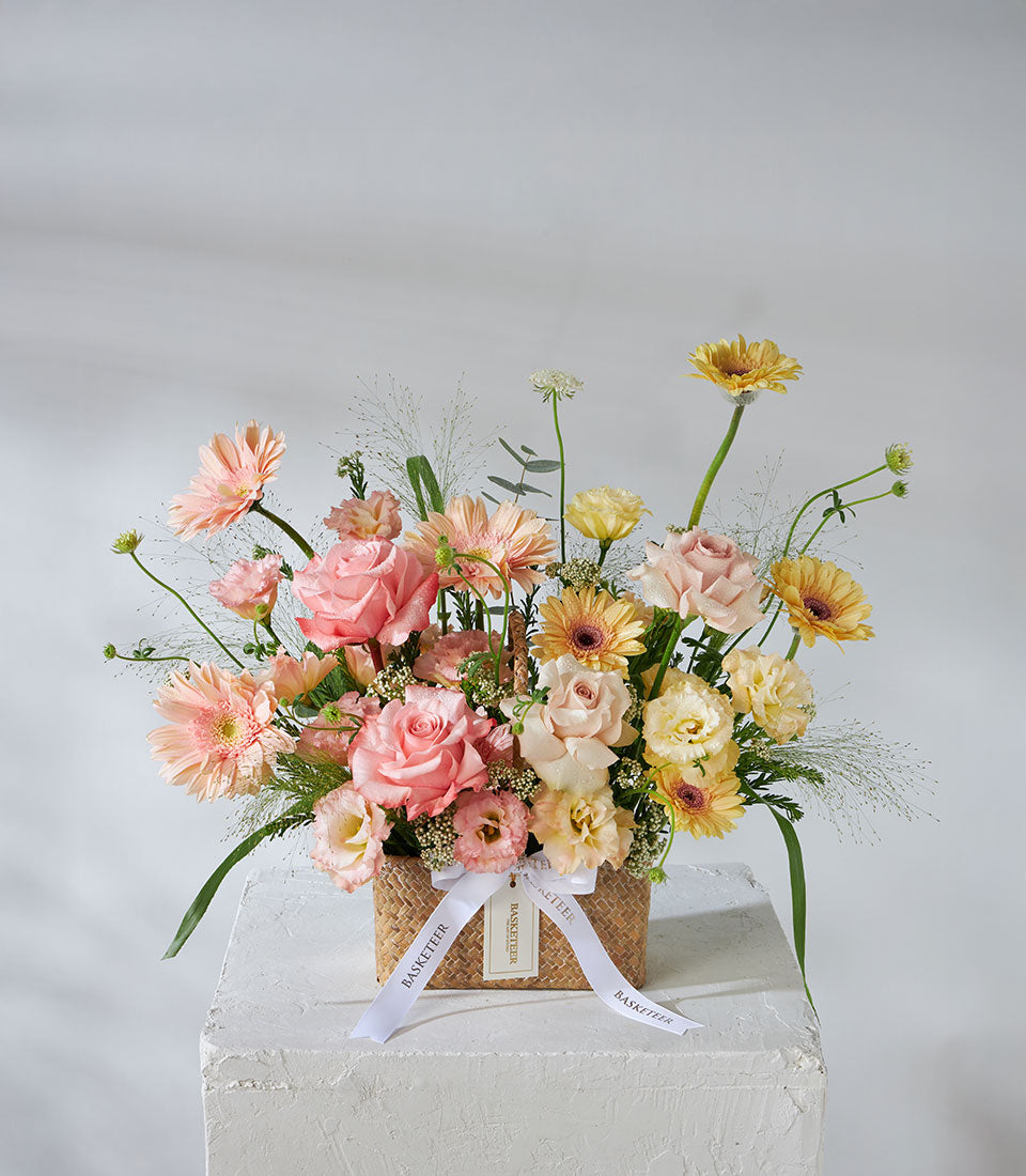 Pastel pink, peach and yellow roses with gerbera daisies in woven basket with white ribbon bow for birthday