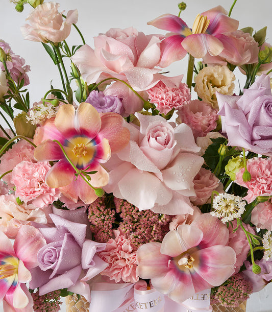 Pastel pink and purple roses with tulips and carnations in a woven flower basket with white ribbon