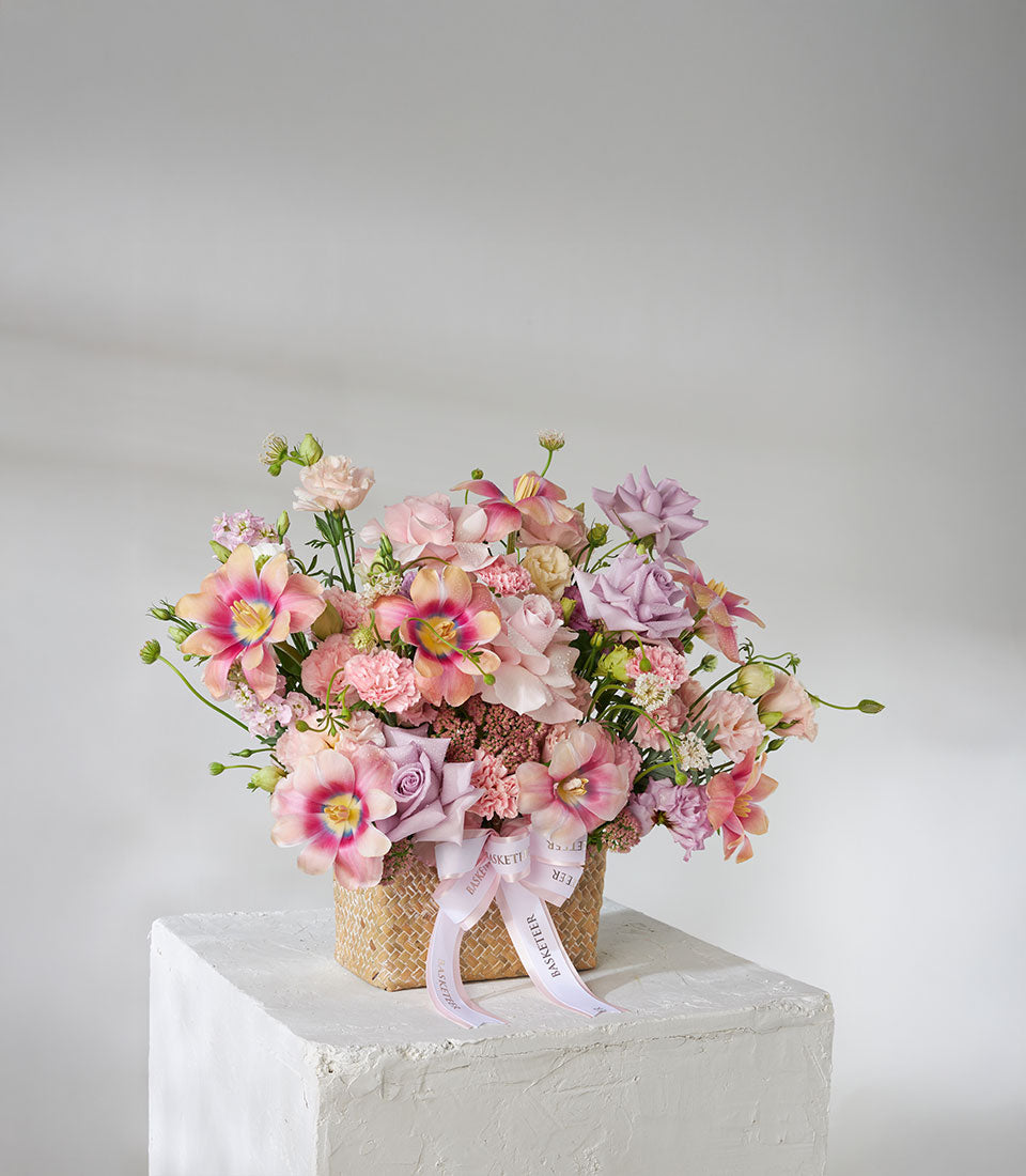 Pastel pink, purple and cream roses with tulips in natural woven basket with pink ribbon
