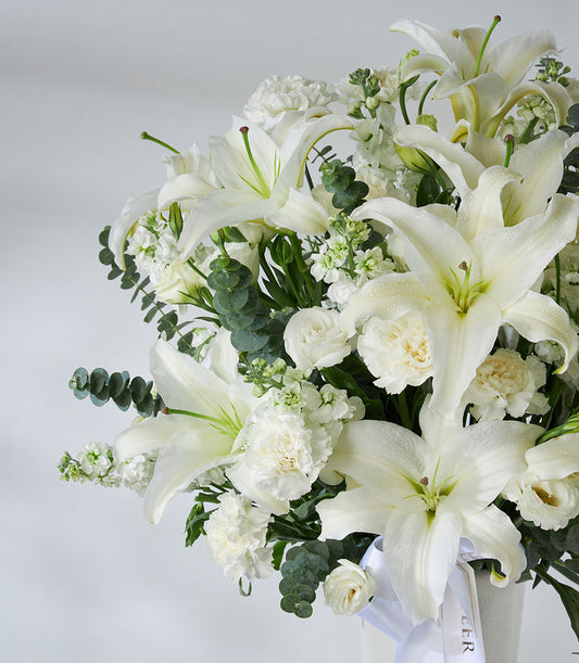 White lilies, carnations, and roses in elegant white vase with eucalyptus and ribbon bow