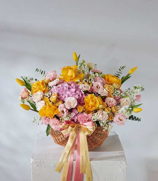 Vibrant yellow roses, pink carnations, and purple hydrangeas in woven wicker basket with gold and pink ribbon bow