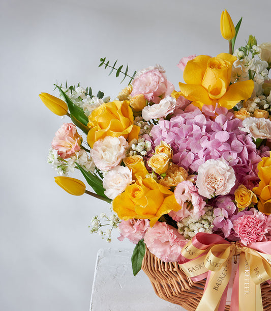 Vibrant yellow roses, pink hydrangeas, and white carnations in woven basket with gold ribbon
