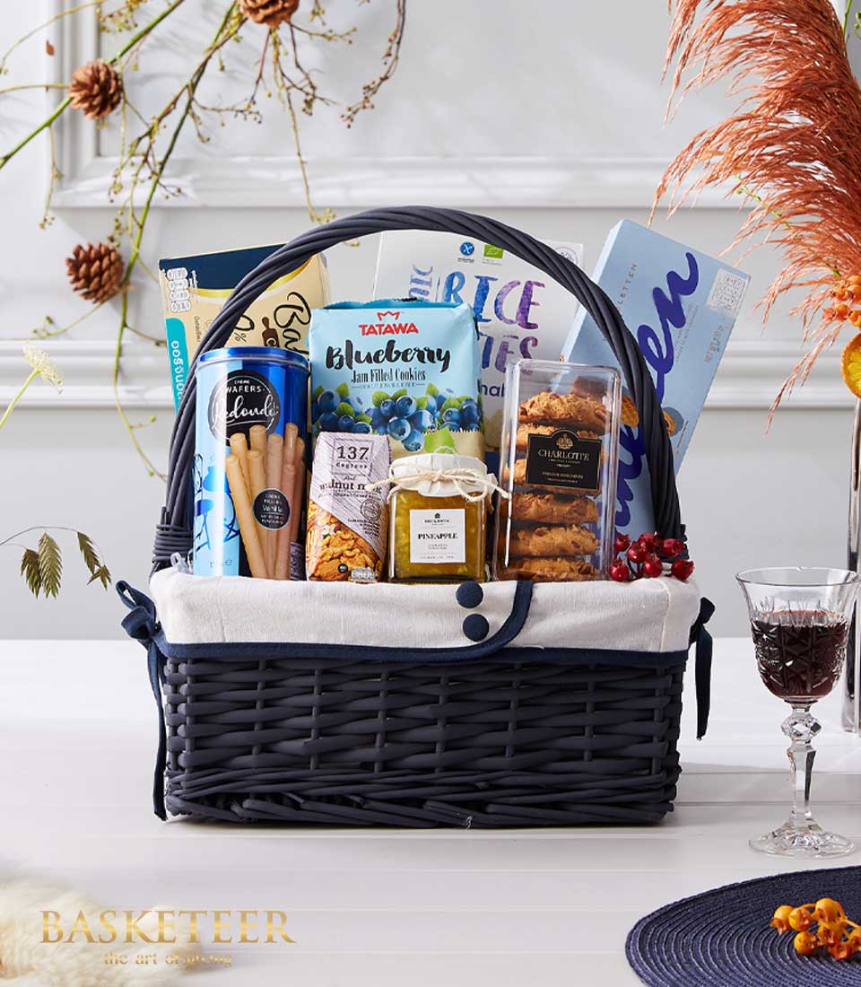 Dark grey wicker gift basket lined with white fabric and blue trim, filled with gourmet treats including jam, wafers, nuts,