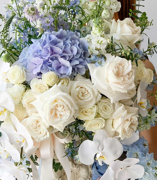 White and blue floral arrangement with hydrangeas, roses, delphiniums and cascading orchids in cream vase