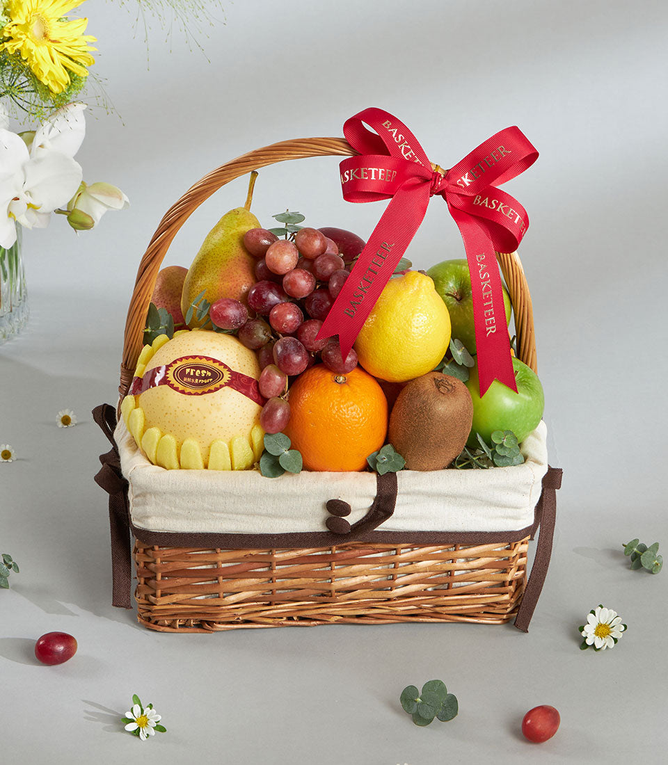 Woven light brown fruit basket with fresh red apples, pears, grapes, and citrus arranged with cream fabric liner and red