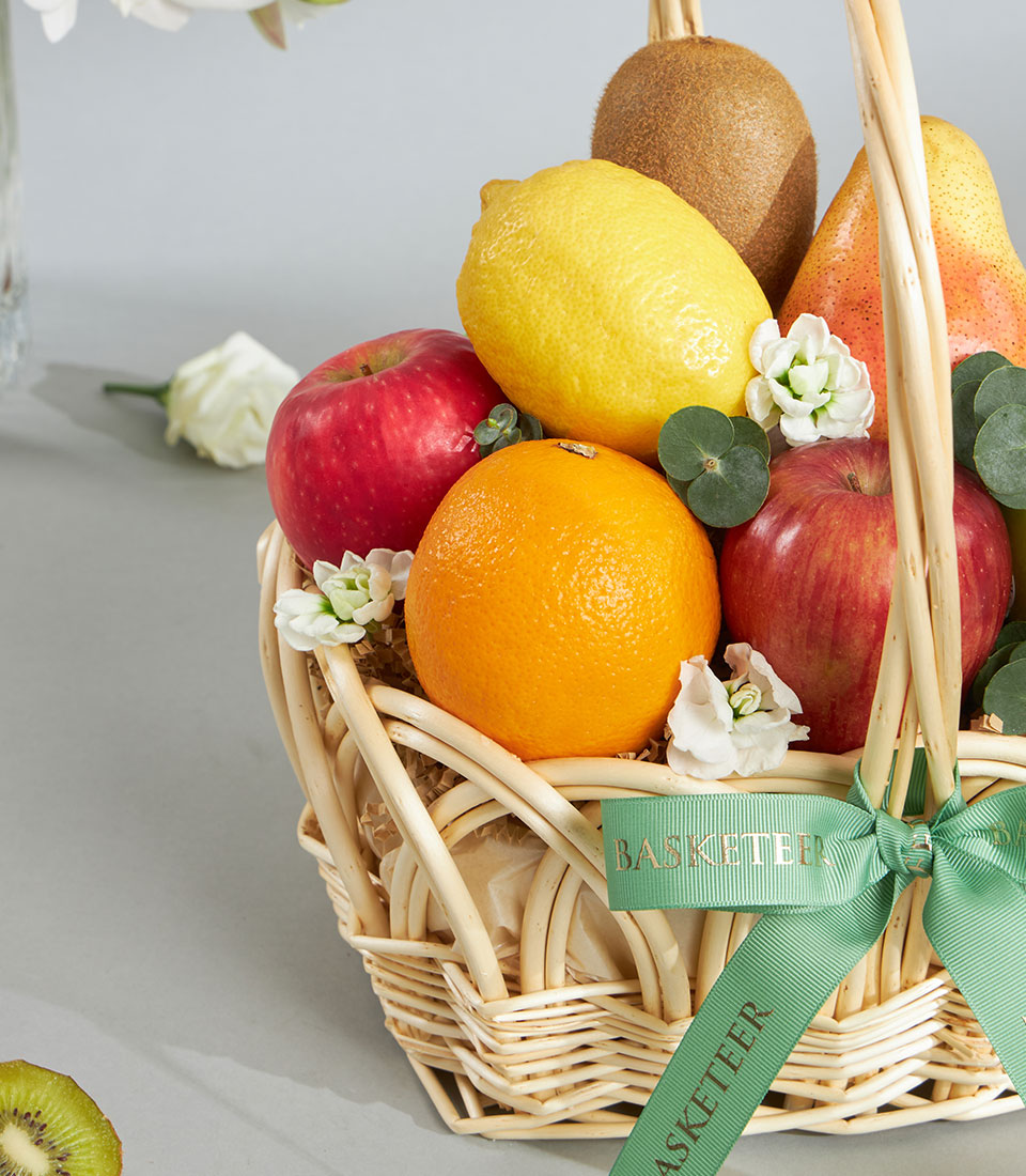 Woven wicker fruit basket with fresh apples, orange, lemon, kiwi, pear, white flowers and green ribbon bow