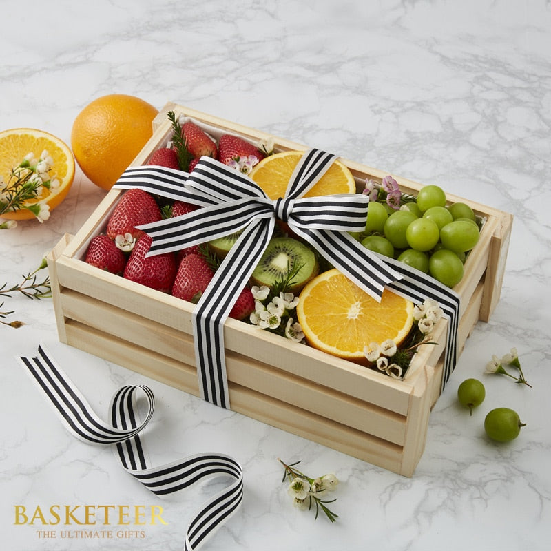 Rustic wooden crate filled with fresh strawberries, oranges, kiwi, and grapes with white flowers and striped ribbon bow, get
