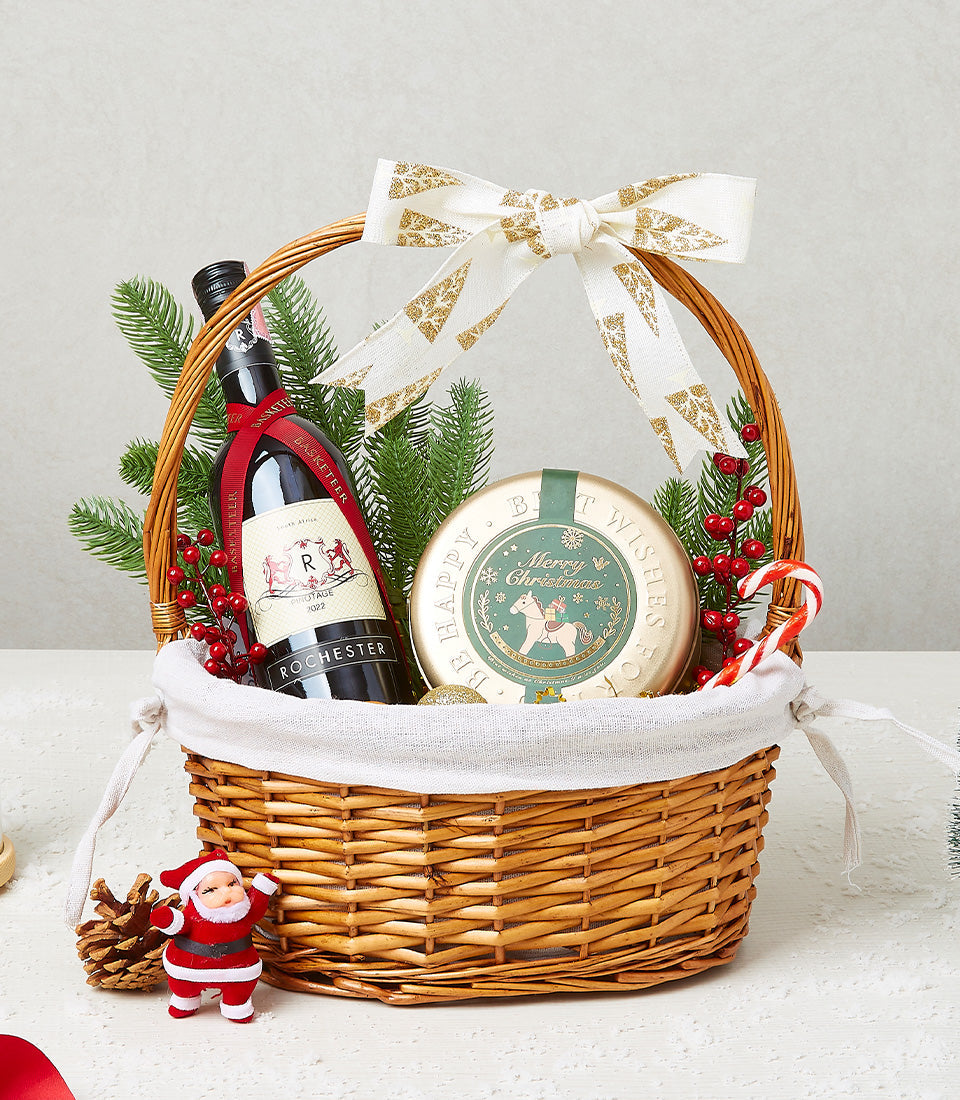 Woven wicker Christmas wine gift basket with red wine, festive gold tin, pine branches, and holiday ribbon bow