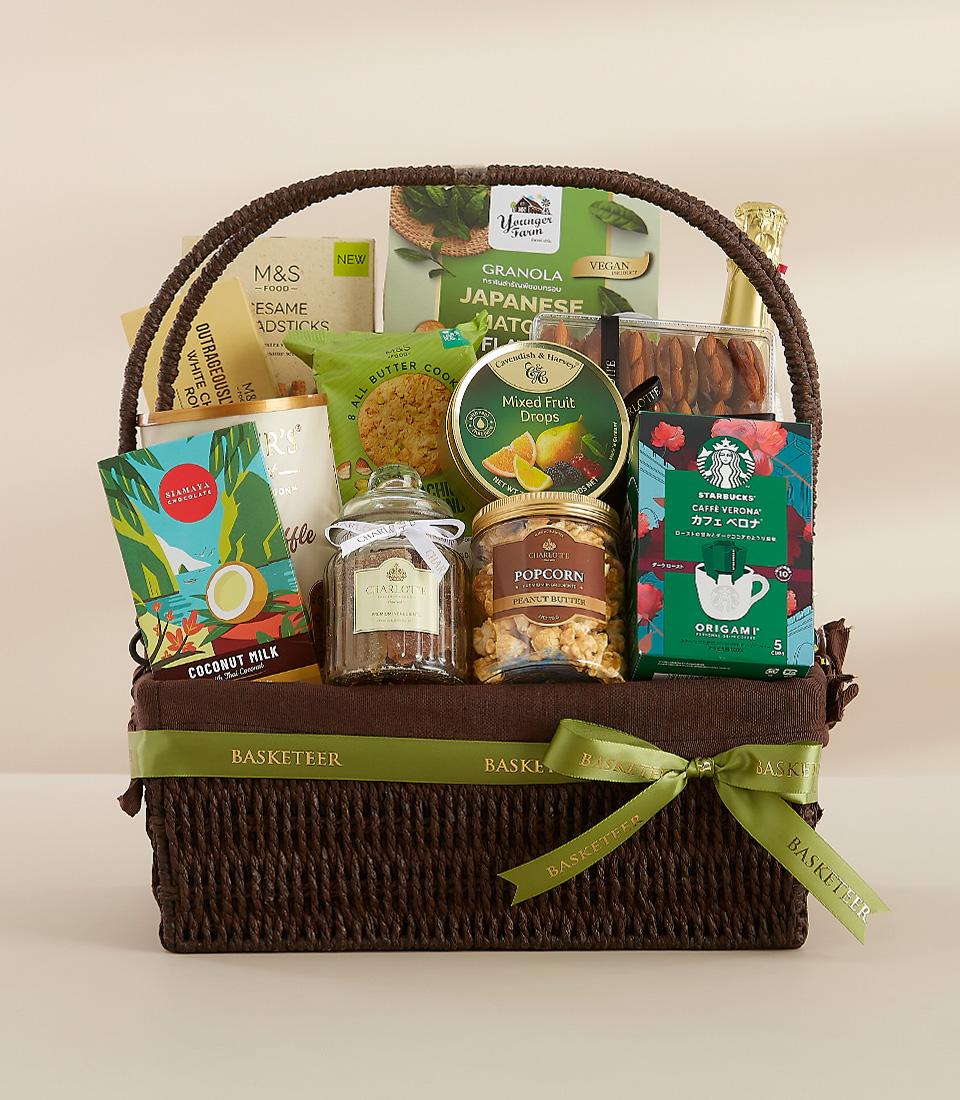 Dark brown woven basket filled with gourmet snacks, matcha granola, coffee, and treats tied with green ribbon, perfect New