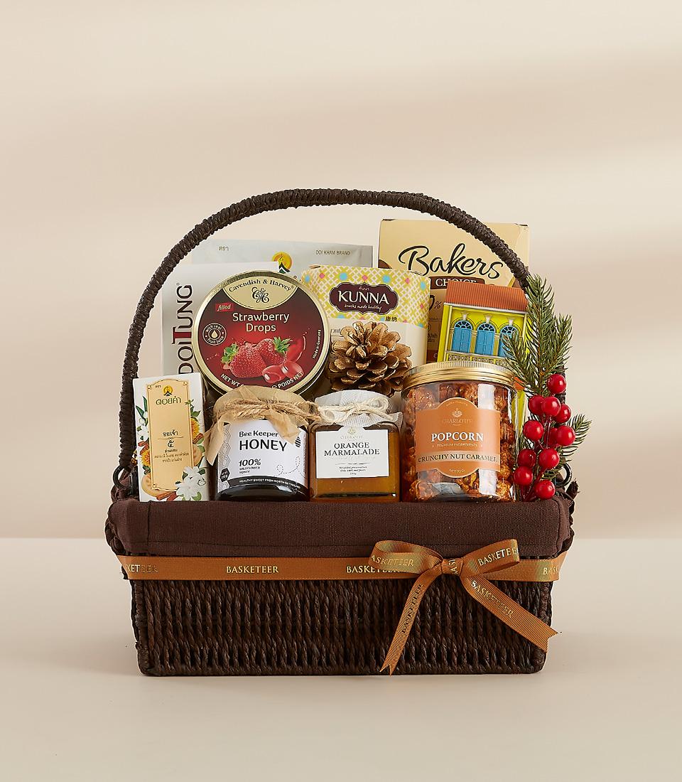 Dark woven gift basket filled with gourmet treats, honey, marmalade, and candies with festive pinecone and red berry accents