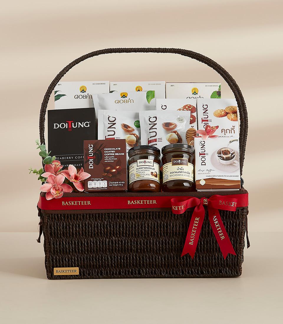 Dark brown woven gift basket with red ribbon filled with Thai gourmet foods, coffee beans, spreads, and fresh pink orchids
