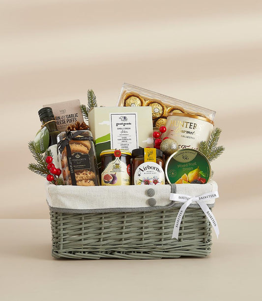 Sage green wicker gift basket with gourmet treats, olive oil, coffee, chocolates, and festive greenery for New Year gifting