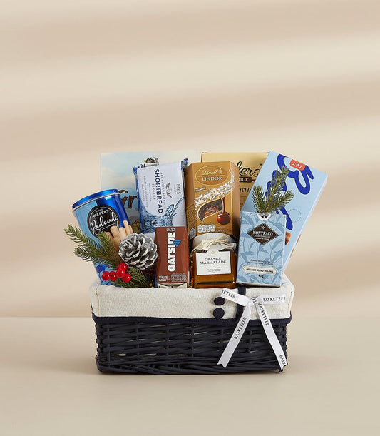 Dark blue wicker wellness hamper with wafers, shortbread, Lindor chocolates, marmalade, oat drink, and tea for New Year