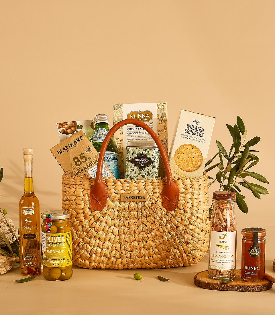 Woven straw gift basket filled with gourmet foods including chocolate, tea, olive oil, crackers, honey, and olives