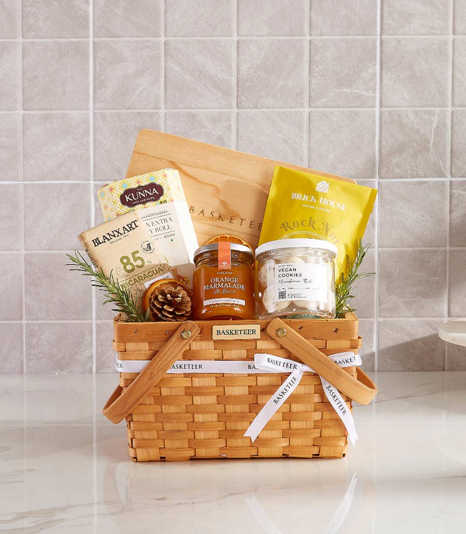 Woven light brown gift basket filled with gourmet foods, cutting board, nuts, marmalade, vegan cookies, and dark chocolate
