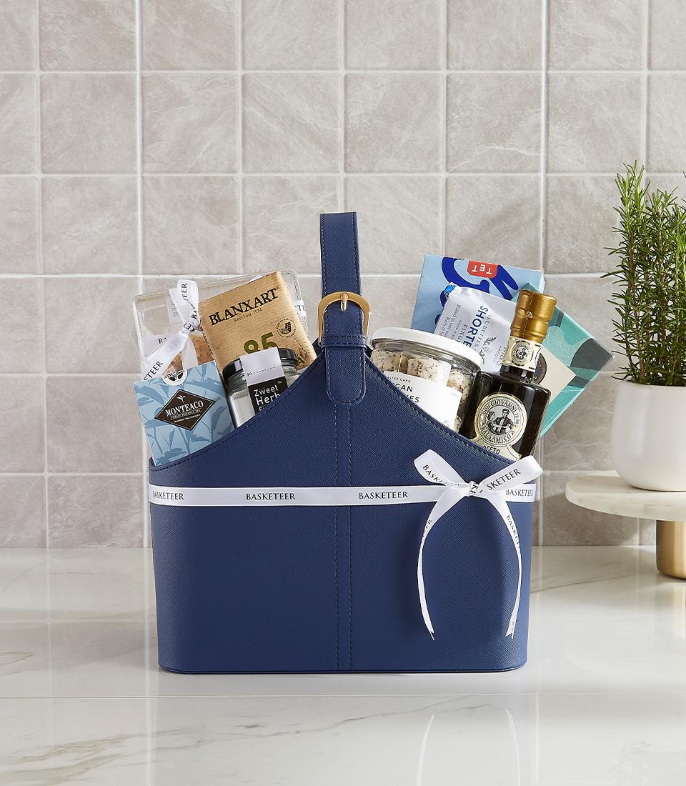 Navy blue leather gift basket with gourmet items, gold buckle handle, and white Basketeer ribbon for New Year gifting