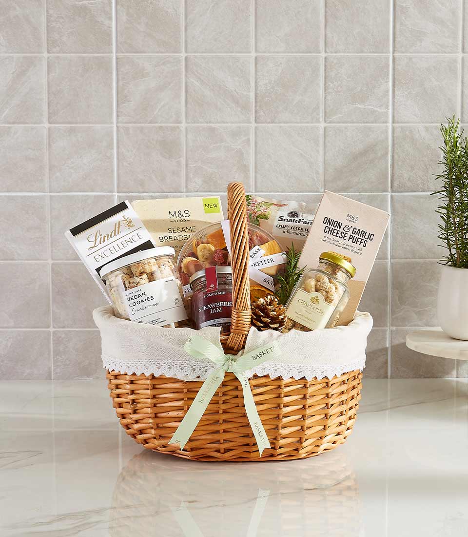 Woven wicker gift basket with gourmet chocolates, jams, nuts, and artisan snacks tied with light green ribbon, perfect for