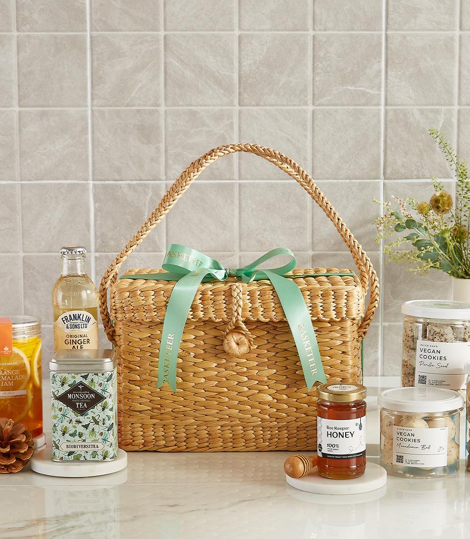 Woven straw gift basket tied with teal ribbon, filled with gourmet tea, honey, vegan cookies, and artisan ginger ale for New
